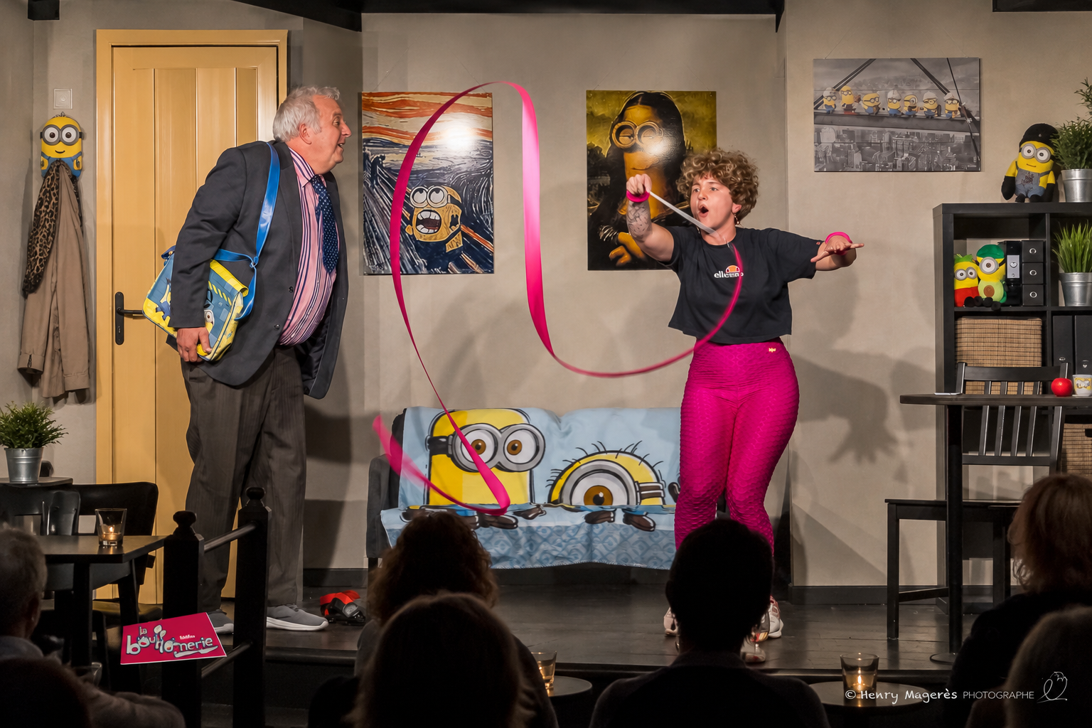 Photo du spectacle à La Bouffonnerie, cadrage large : deux interprètes sur scène avec un ruban rose et le public au premier plan. © Henry Magerès Photographe.
